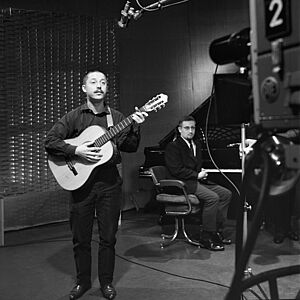 Der junge Wolf Biermann steht mit Gitarre in einem Aufnahmestudio und singt, im Hintergrund sitzt ein Mann auf einem Klavierhocker